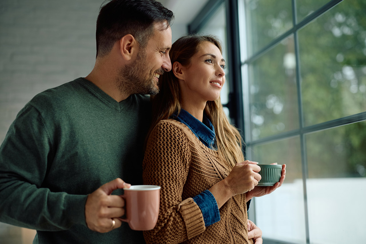 Smiling woman day dreaming while drinking coffee with her husband by the window at home